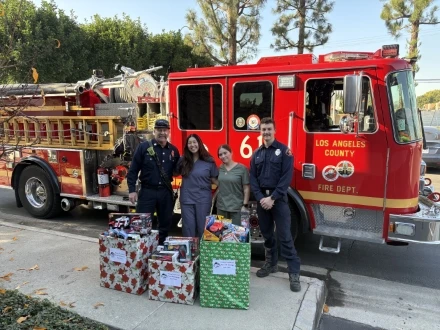 Firefighters and dental staff smiling beside holiday toy donation boxes in front of a Los Angeles County fire truck.