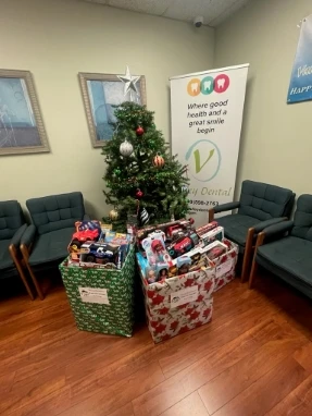 Toy-filled holiday gift boxes and bags around a Christmas tree in a dental office waiting area.